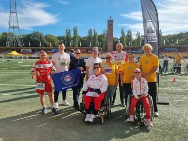 Grupa osób, w tym dwie na wózkach inwalidzkich, pozuje do zdjęcia na bieżni stadionu lekkoatletycznego podczas imprezy sportowej.
